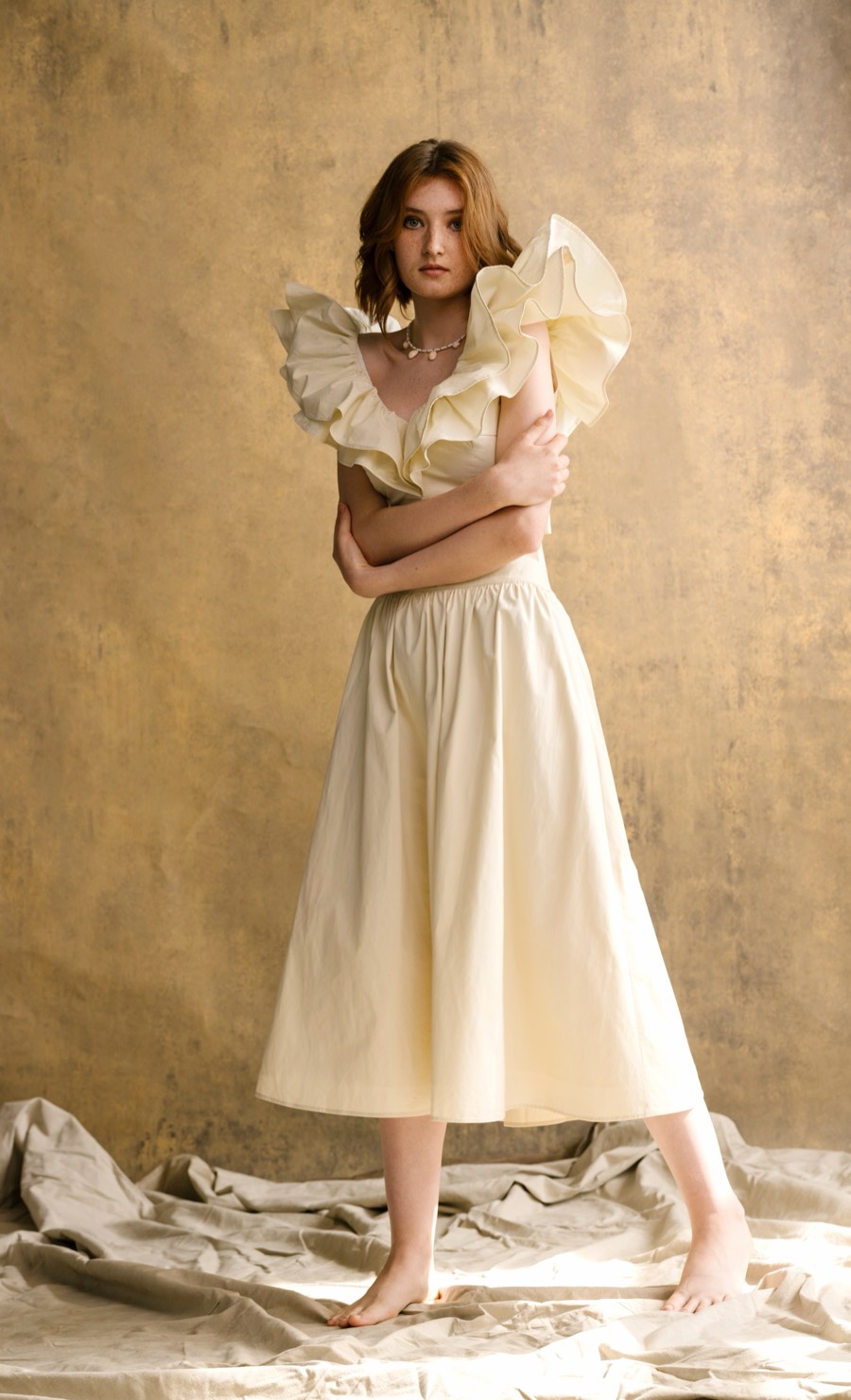A model in a cream Minem ruffle top and full midi skirt, standing barefoot on draped linen in front of a gold backdrop.